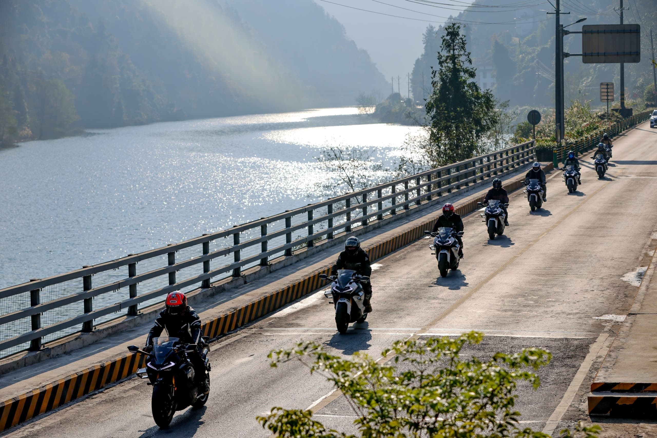 Motorcyclists in a line ride on a bridge next to a wide, sun-dappled river.