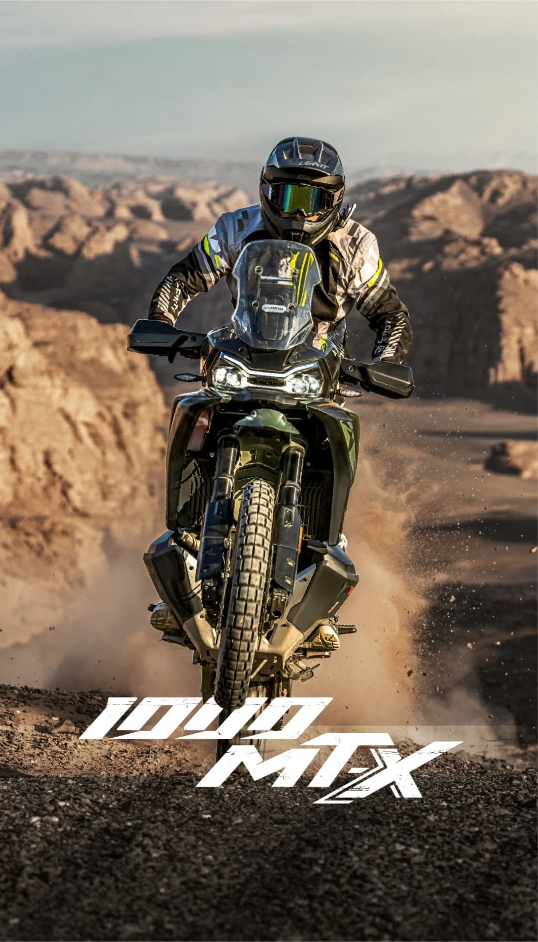 A rider on a dark adventure motorcycle kicks up dust in a rocky desert landscape.