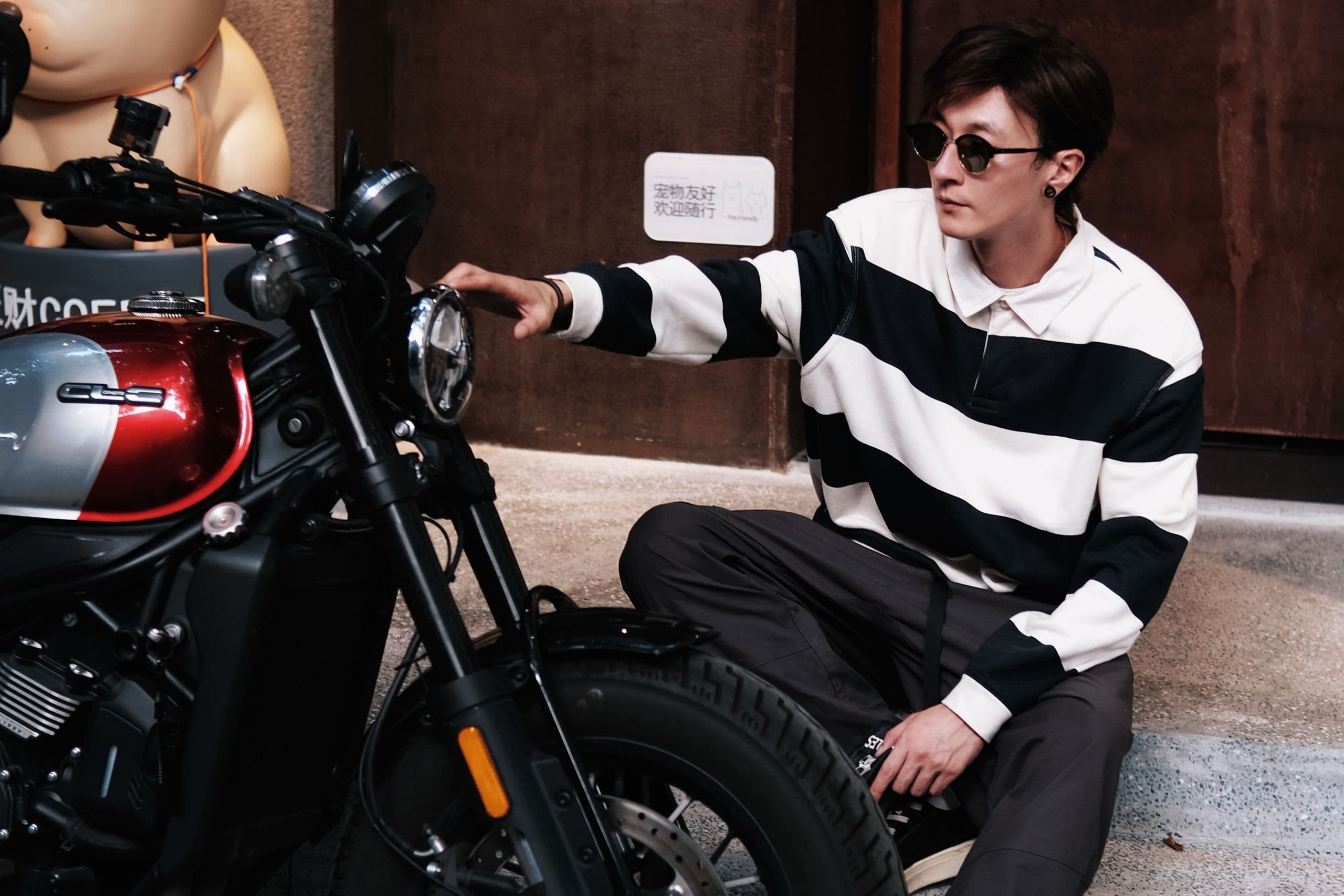 Man in striped shirt sits casually on steps beside a black motorcycle.