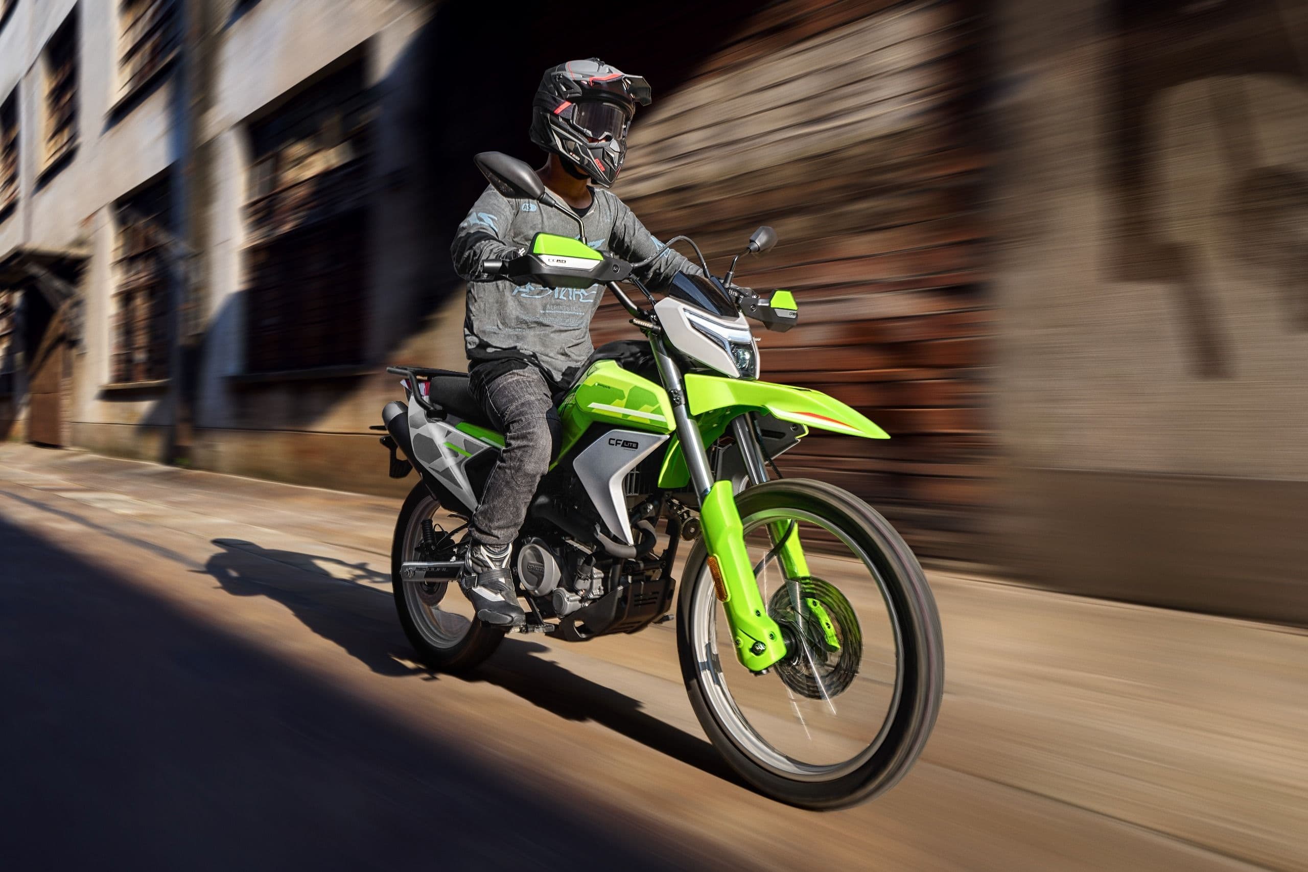 Rider in helmet and gear on a green dirt bike speeding through a city alley.