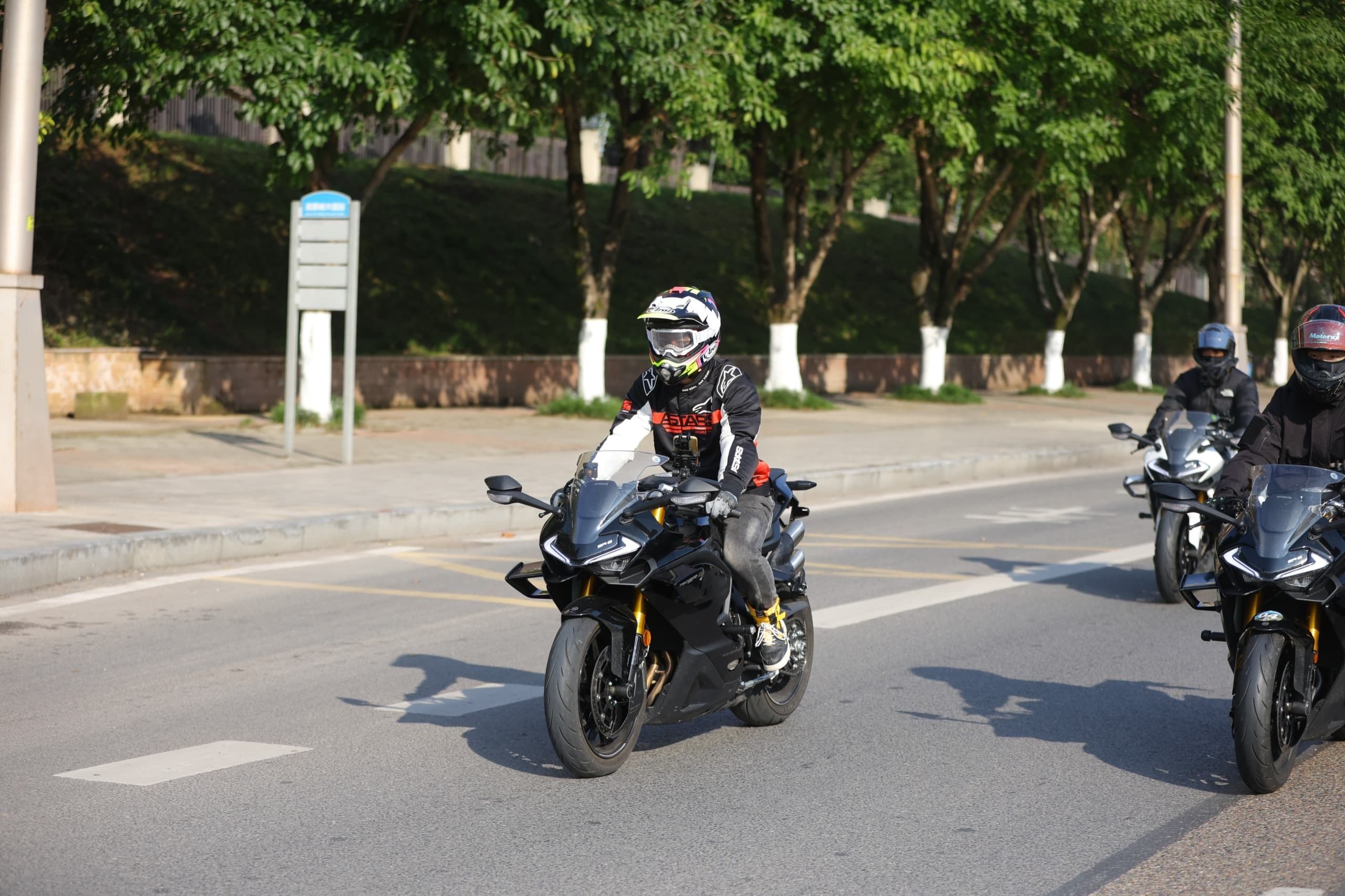 Two riders on black sport motorcycles travel on a tree-lined street.