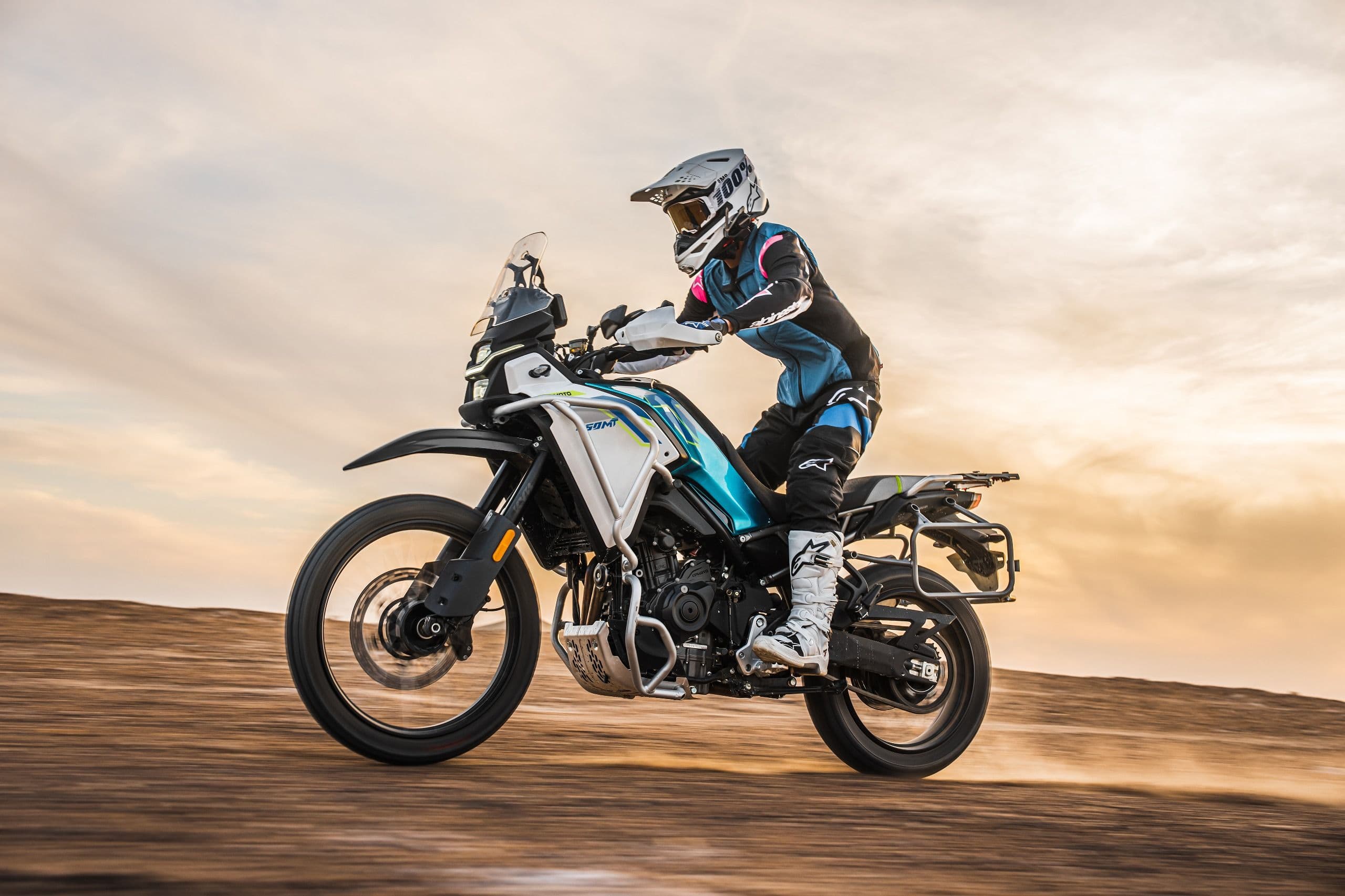 Rider in protective gear on a white and blue adventure motorcycle in a dusty desert landscape.