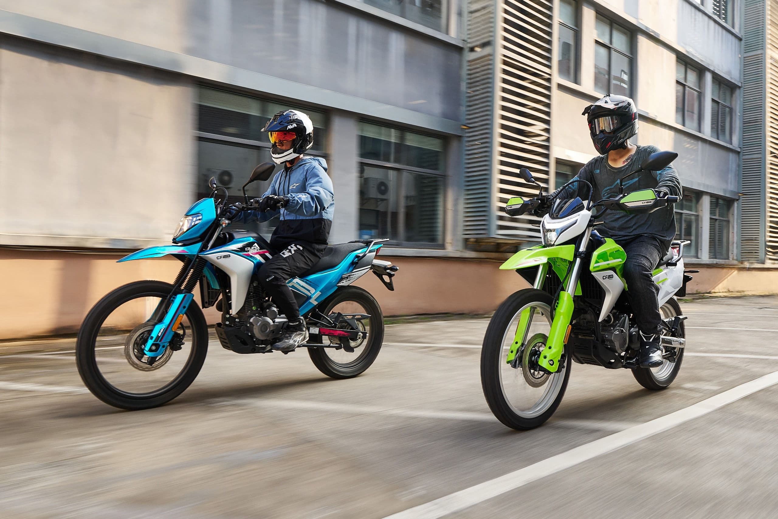 Two riders on a blue and a green dirt bike parked on an asphalt lot.