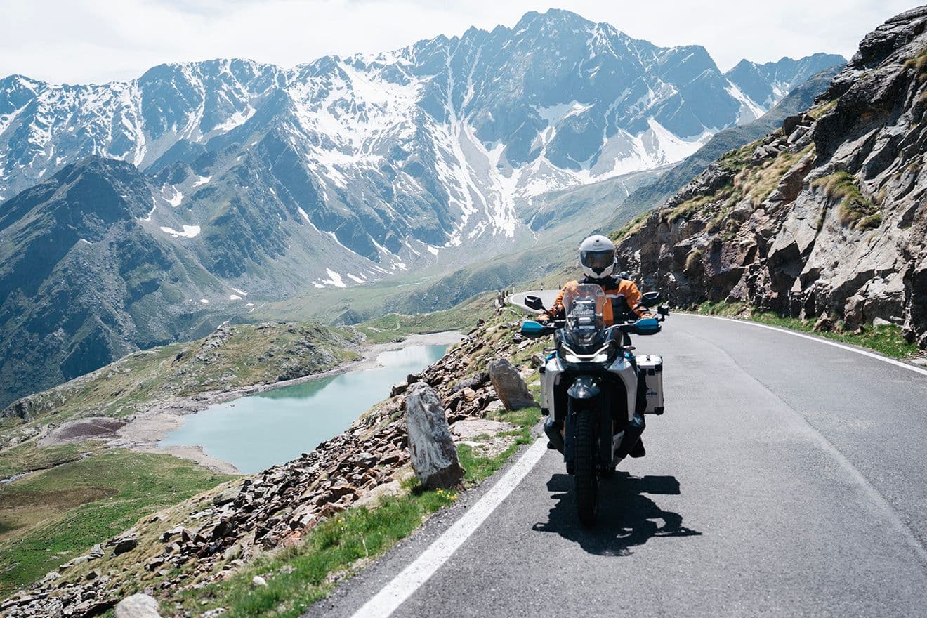 Motorcyclist on a winding mountain road with snow-capped peaks and a turquoise lake.